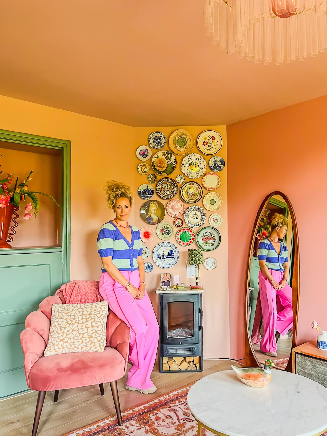 Interieurstylist Puck Steijling in een woonkamer met okergele wanden, een verzameling vintage wandborden boven een houtkachel en een oudroze fauteuil